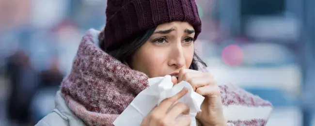 A woman, wearing a winter hat and scarf, holds a tissue while coughing.